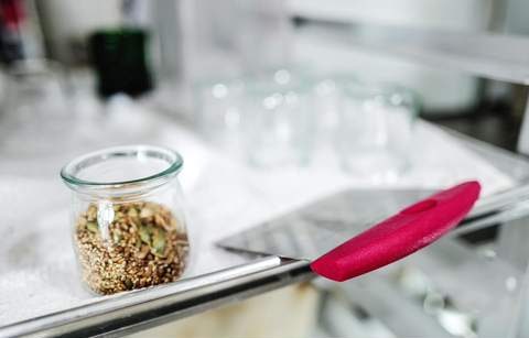 A glass jar of seeds on the flour-dusted bench, beside a red dough scraper